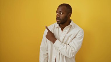 Ciddi ifadeleri olan genç bir adam, izole edilmiş sarı bir arkaplanı işaret ediyor, odaklanmasını ve kararlılığını vurguluyor..