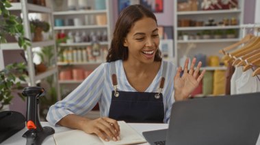 Dekorasyonlu bir mağazada dizüstü bilgisayarla çalışan genç bir kadın dükkan sahibi, görsel çağrı sırasında gülümsüyor ve el sallıyor, etrafı dekorasyon malzemeleriyle çevrili..