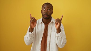 Beyaz tişörtlü yakışıklı Afro-Amerikan adam izole edilmiş sarı arka planda parmaklarını yukarı kaldırıyor.