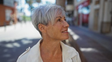 Güzel gri saçlı kadın, kısa saçlı güneşli bir şehir caddesinde yürüyor, gülümsüyor ve şehir atmosferinin tadını çıkarıyor..