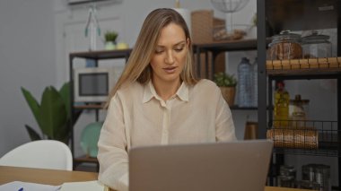 Modern ofis ortamında çalışan bir kadın, sarışın ve dizüstü bilgisayara odaklanmış, iç mekan bitkileri ve şık bir iç mekan ile çevrili, çağdaş bir çalışma alanını yansıtıyor..