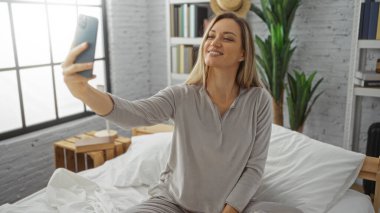 Parlak yatak odasında selfie çeken genç bir kadın bitkiler ve kitaplarla yatakta rahat ve rahat bir yaşam tarzı sergiliyor. Modern dekorasyon ve kişisel alan..