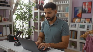 Ev dekorasyonu mağazasında dizüstü bilgisayarda çalışan genç İspanyol bir adam. Etrafı bitkiler, raflar ve dekoratif malzemelerle çevrili.