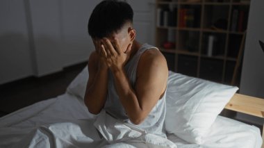 Genç bir adam, elleri loş ışıklı bir yatak odasında, bir yatakta oturuyor, gergin ya da üzgün görünüyor, rahat bir ev atmosferiyle çevrili..