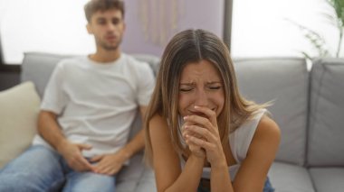 Oturma odasındaki kanepede ağlayan kadın, arka planda erkek, evde duygusal bir çift durumu tasvir ediyor..