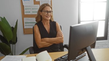 Modern ofis ortamında gülümseyen bir kadın, kollarını kavuşturup bilgisayar başında oturuyor, parlak, kapalı bir ortamda profesyonelliği ve güveni iletiyor..