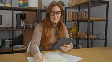 Bir ofis ortamında çalışan kadın, tablet ve notlara odaklanmış, mutfak aletleri ve dekorlarla çevrili, rahat bir iş ortamı yaratıyor..
