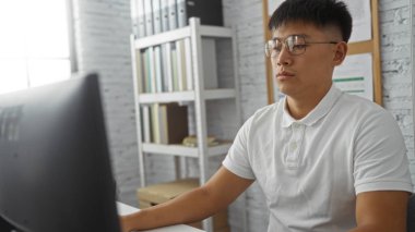 Genç adam, arka planda kitap ve belgelerle dolu bir ofiste bilgisayarla çalışıyor.