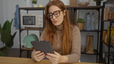 Ofiste çalışan bir kadın kırmızı saçlı ve gözlüklü bir tablet okuyor. Kapalı alanda rahat bir süveter giyiyor. Raflar ve ev mutfağına bitişik dekor yerleştiriyor..