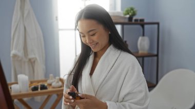 Genç Çinli bir kadın spa ortamında, beyaz bir bornoz giyiyor, elinde bir cilt bakım merkezinde ürünü tutarken gülümsüyor.