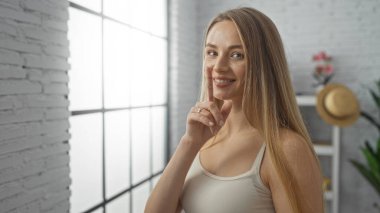 Kadın sessiz hareketler yapıyor, parmakları dudaklarında, kapalı bir odada gülümsüyor, aydınlık, modern bir oturma odasında ve beyaz tuğladan bir arka planı var..