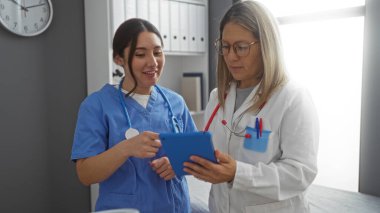 Hastane odasında iş birliği yapan doktorlar, kapalı klinik ortamındaki bir tablet üzerinde hasta bilgilerini inceleyen iki kadın, ekip çalışması ve profesyonel sağlık hizmetleri bağlamını gözler önüne seren.
