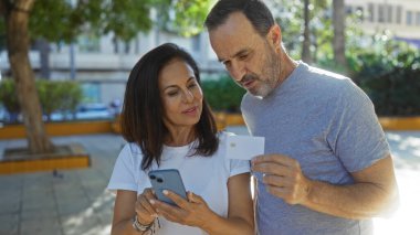 Şehir parkında ayakta duran orta yaşlı İspanyol çift ellerinde akıllı telefon ve kart, aşkı, aile bağını ve modern teknolojiyi temsil ediyorlar..