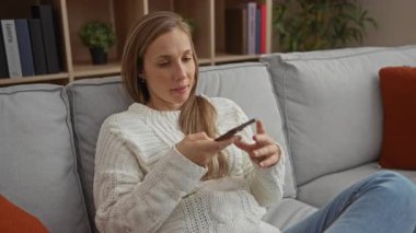 Oturma odasındaki kanepede oturan kadın akıllı telefon ses komutuyla ev ortamında rahat bir süveter giyiyor..