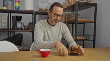 Ofisteki İspanyol bir adam, arka planda rafları ve kırmızı bardağı olan ahşap bir masada otururken akıllı telefonuna bakıyor..