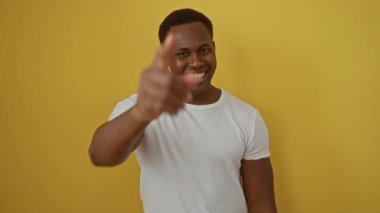 Sarı arka planda gülümseyen genç adam kameraya beyaz tişörtlü neşeli bir ifadeyle bakıyor.