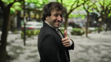 Ağaçlarla ve park edilmiş arabalarla dolu bir caddede baş parmağıyla dışarıda gülümseyen bir adam..