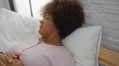 Yatakta kıvırcık saçlı bir kadın modern bir yatak odasında farklı duygular sergiliyor. Tarafsız bir arkaplanda rahatlama ve tefekkür sergiliyor..
