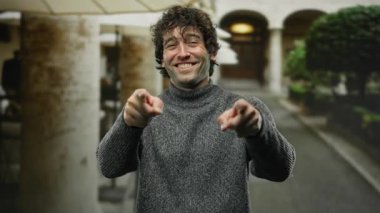 Gülümseyen ve el kol hareketi yapan genç bir adam bir şehir caddesinde dikilirken, bir şehir ortamında güven ve dostluk yayarak buraya gelir..