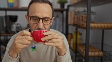 Kadehler takan olgun bir adam, arka planda raflar ve ev eşyaları olan rahat bir mutfakta kırmızı bardaktan çay içiyor..