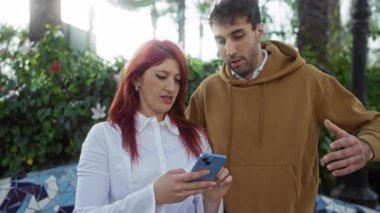 Kadın ve erkek açık hava parkında yan yana durmuş yeşillik ve mozaik sanat eserleriyle çevrili akıllı telefon etkileşimlerini paylaşıyorlar..