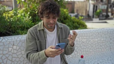 Şehir parkında telefonla kutlama yapan genç adam, yeşil ve mozaik banklarla çevrili, çapraz parmaklar ve neşeli bir ifadeyle heyecan gösteriyor..