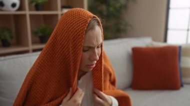 Turuncu bir battaniyeye sarılmış bir kadın, rahat bir oturma odasında kanepede otururken keyifsiz ve dalgın görünüyor. Bu da içeride bir yalnızlık ve yansıma anını işaret ediyor..