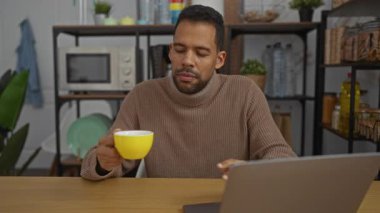 Ofisteki genç adam laptopun önünde sarı bardaktan kahve içiyor. Arka planda modern mutfak rafları var. Odaklanmış ve rahat görünüyor..