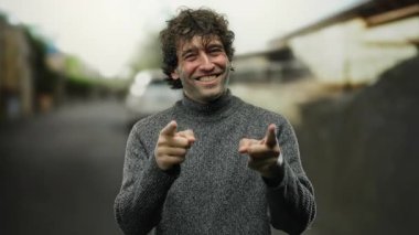 Şehir sokağında gülümseyen genç bir adam güvenli bir şekilde süveter giyiyor neşeli ve çekici bir hava sergiliyor hafif ışıklandırma altında bulanık şehir arkaplanı ile.