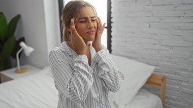 Başı ağrıyan bir kadın, parlak bir yatak odasında, bir yatakta otururken, çizgili pijama giyiyor, sakin bir evde oturmaktan rahatsız olduğunu söylüyor..