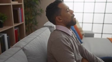 Genç adam rahat bir oturma odasında kanepede dinleniyor. Yumuşak yastıklı ve rahatlatıcı bir çevresi olan huzurlu bir iç mekan atmosferi sergiliyor..