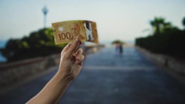 Kanadalı banknot tutan adam, açık gökyüzü ve arka planda ağaçlarla şehir ortamında finans kavramına odaklanıyor..