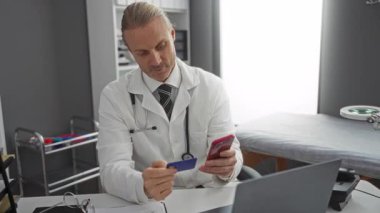 Klinikteki doktor online ödeme için telefon kullanıyor, kart tutuyor ve tıbbi büronun iç kısmında masa başında oturuyor..