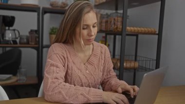 Ofiste dizüstü bilgisayar ve telefonla çalışan sarışın bir kadın, süveter giyiyor, yüz ifadelerini gösteriyor, raflarla çevrili, rahat ve verimli bir iş ortamı olduğunu gösteriyor..