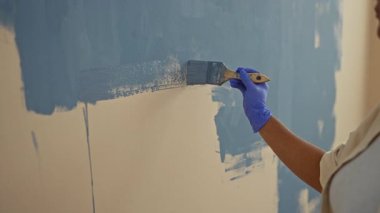 Evdeki duvarları fırça ve eldivenlerle maviye boyayan kadın, modern oturma odasındaki loş bir projeye işaret ediyor..