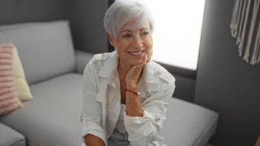 Güzel, gri saçlı, kısa saçlı bir kadın oturma odasında gülümsüyor, huzurlu ve neşeli bir ifade sergiliyor, modern dekorlu rahat bir ev ortamında..