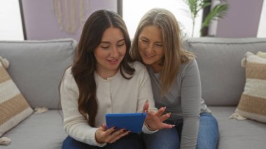Anne ve kızı sıcak bir oturma odasında birbirlerine bağlanıyorlar, bir tablet kullanarak neşeli bir anı paylaşıyorlar, aile ortamlarında sevgi ve sıcaklığı ifade ediyorlar..