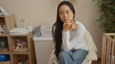 Genç Çinli kadın bebek oyuncakları ve mobilyalarla dolu rahat bir yatak odasında düşünceli bir şekilde oturuyor. Huzurlu ve rahat bir iç mekan izlenimi veriyor.