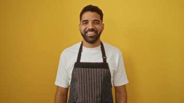 Yakışıklı, genç, İspanyol bir adam. Önlüğüyle güvenle gülümsüyor. Kalın sarı bir arka planı var.