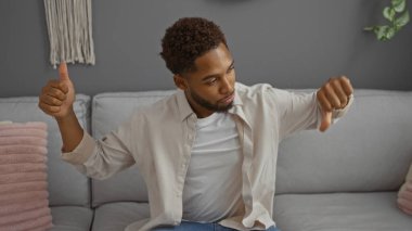 Genç bir adam, modern dekorlu rahat bir oturma odasında kanepede otururken baş parmak hareketi yapıyor.