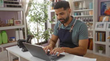 Dizüstü bilgisayarda çalışan genç bir İspanyol, arka planda raflar dolusu ürünle birlikte bir ev dekorasyonu dükkanında çalışıyor.