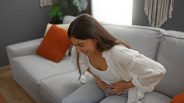 Modern bir oturma odasında rahat bir koltukta oturan karın ağrısı çeken bir kadın karnını tutarken kederli bir ifade sergiliyor.