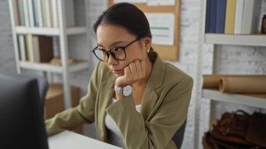 Ofiste çalışan genç Çinli bir kadın, bilgisayara odaklanmış, arka planda kitap rafları ve mantar panosu olan, gözlük takan ve iş kıyafeti giyen..