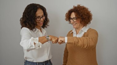 İki olgun İspanyol kadın gülümsüyor ve yumruk tokuşturuyor, soyutlanmış beyaz bir arka plana karşı neşeli bir dostluk sergiliyorlar, günlük kıyafetler ve gözlük takıyorlar..