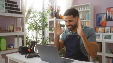 Elleri havada genç bir adam, bir ev dekorasyonu mağazasında kutlama yapıyor, önlük giyiyor ve elinde dizüstü bilgisayar ve tarayıcı ile tezgahın arkasında duruyor, çeşitli dekoratif eşyalar ve bitkilerle çevrili.