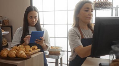 Pastanede pastalarla çalışan, tablet ve bilgisayar kullanan, önlük takan, görevlere odaklanan, kapalı alandaki çekimlere odaklanmış kadınlar.