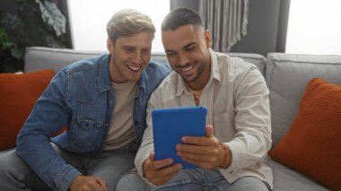 Erkek arkadaşlar, oturma odasında oturmuş tablete bakarken gülümseyen yakışıklı yetişkinler.