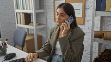 Ofiste, rafları ve evrakları olan bir bilgisayarın yanında otururken akıllı telefondan konuşan genç bir kadın.