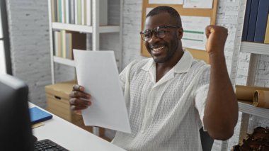 Genç, Afro-Afrikalı, Amerikalı adam ofiste evrak okurken başarıyı kutluyor, arka planda kitaplıkların olduğu olumlu ve üretken bir iş ortamı sergiliyor..