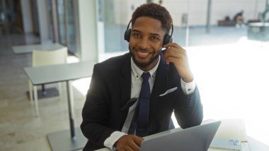 Modern ofis ortamında çalışan, kulaklık takan, laptopunun önünde güvenle gülümseyen genç bir adam profesyonel ve üretken bir çalışma ortamı öneriyor..